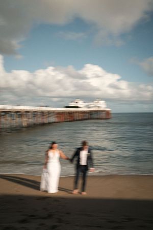 Blurred artistic photo of a couple holding hands on a beach with a pier and cloudy sky.