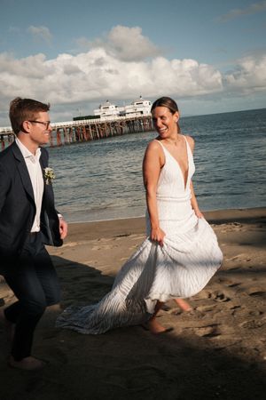 Newlyweds having fun on sandy beach with pier in background during sunset photoshoot.