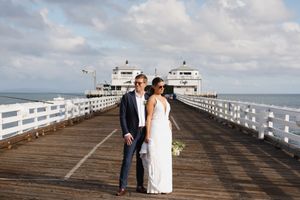 A couple in wedding attire poses on a wooden pier with the ocean and cloudy sky in the background.