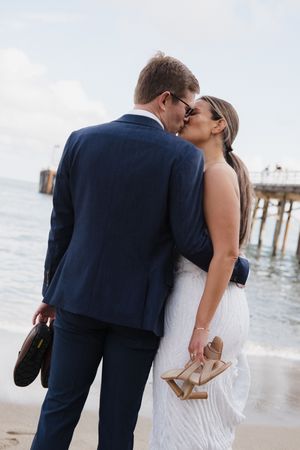 A couple in wedding attire shares romantic moments walking along a beach with a pier in the background.