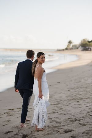 A couple in wedding attire walks barefoot along a sandy beach at sunset.
