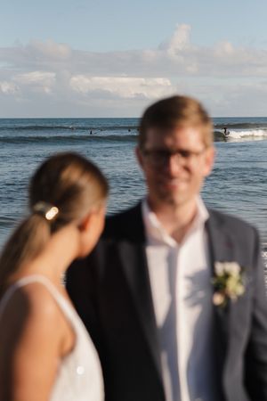 A romantic beach wedding portrait with ocean waves and surfers visible in the background.