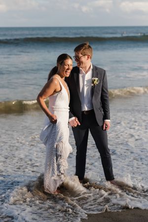 A joyful wedding couple stands in the surf as waves crash around their feet at the beach.