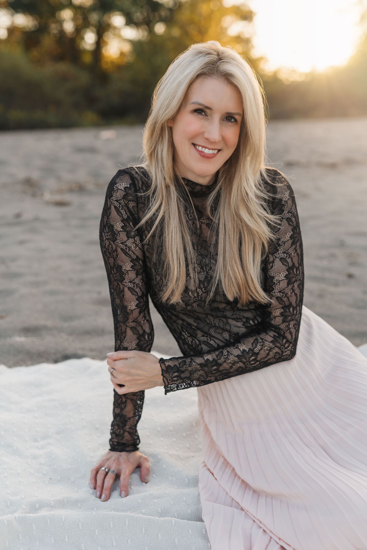 Michelle Hugill seated on a sandy shoreline at sunset, wearing a black lace top and soft pink skirt