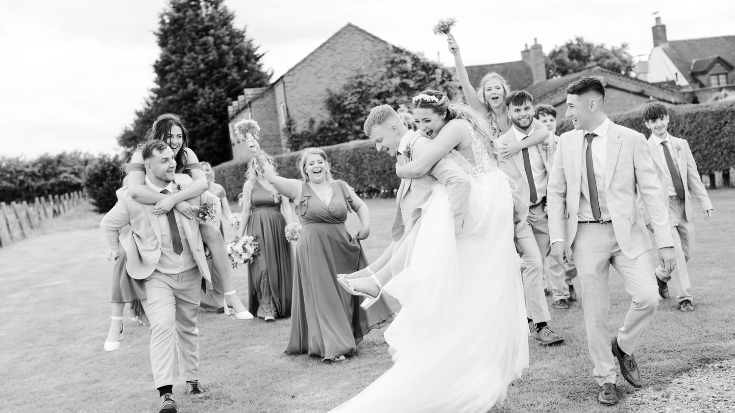 Black and white photo of a large bridal party cheering, shouting and giving piggy backs at Oak Farm Hotel, staffordshire