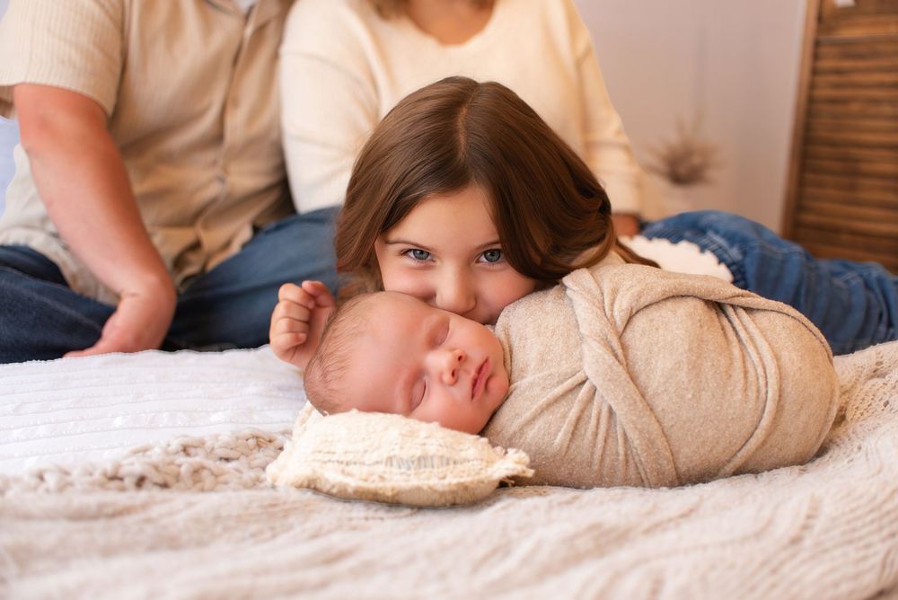 Young girl with dark hair rests her chin on a swaddled newborn baby while parents sit behind them on a bed.