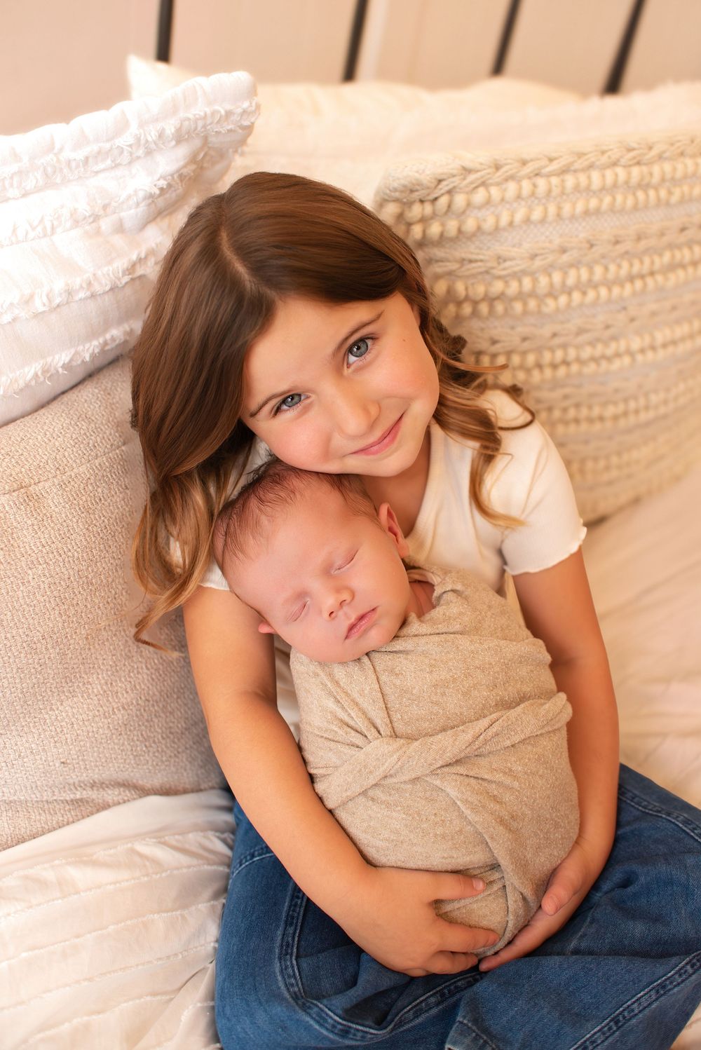 Young girl with brown hair holds sleeping swaddled newborn baby sibling while sitting on a bed with pillows.