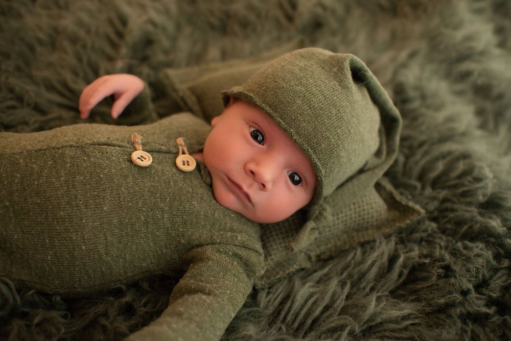 Newborn baby in green knit outfit and hat lying on fluffy gray fur blanket.
