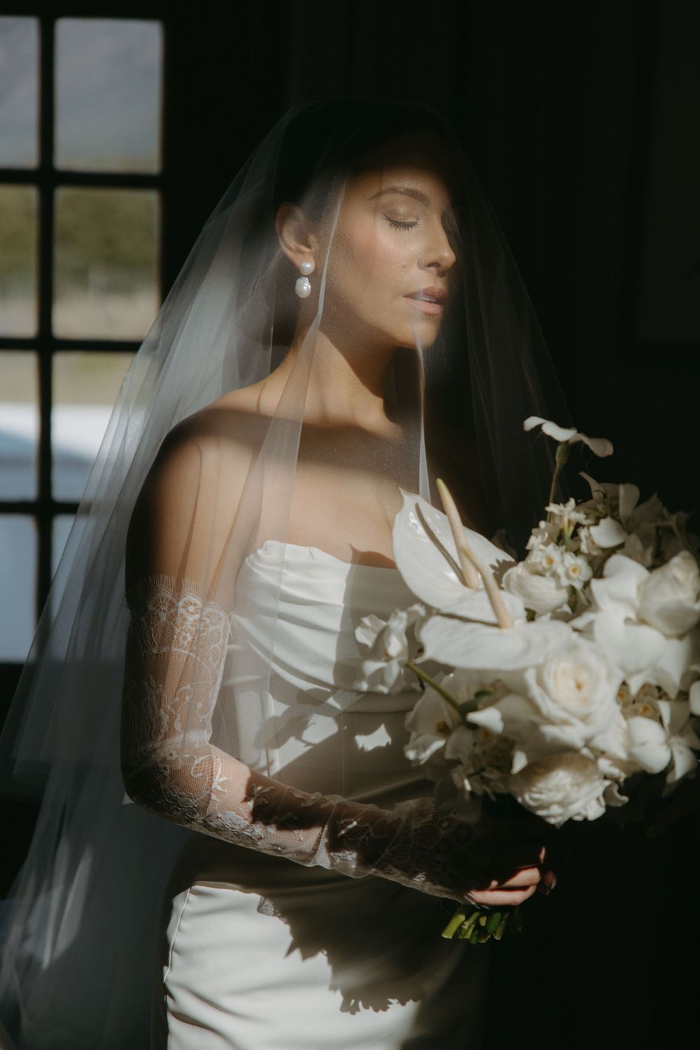 Elegant bridal portrait with white bouquet and veil in dramatic dark lighting.