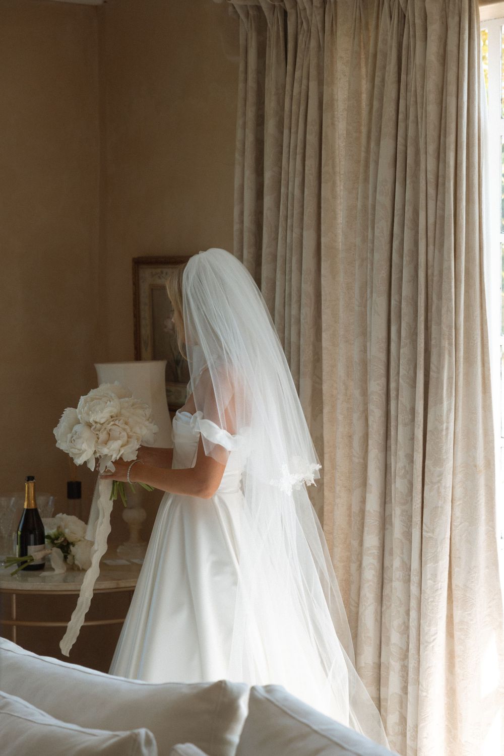 A bride in a classic white wedding gown stands near curtained windows.
