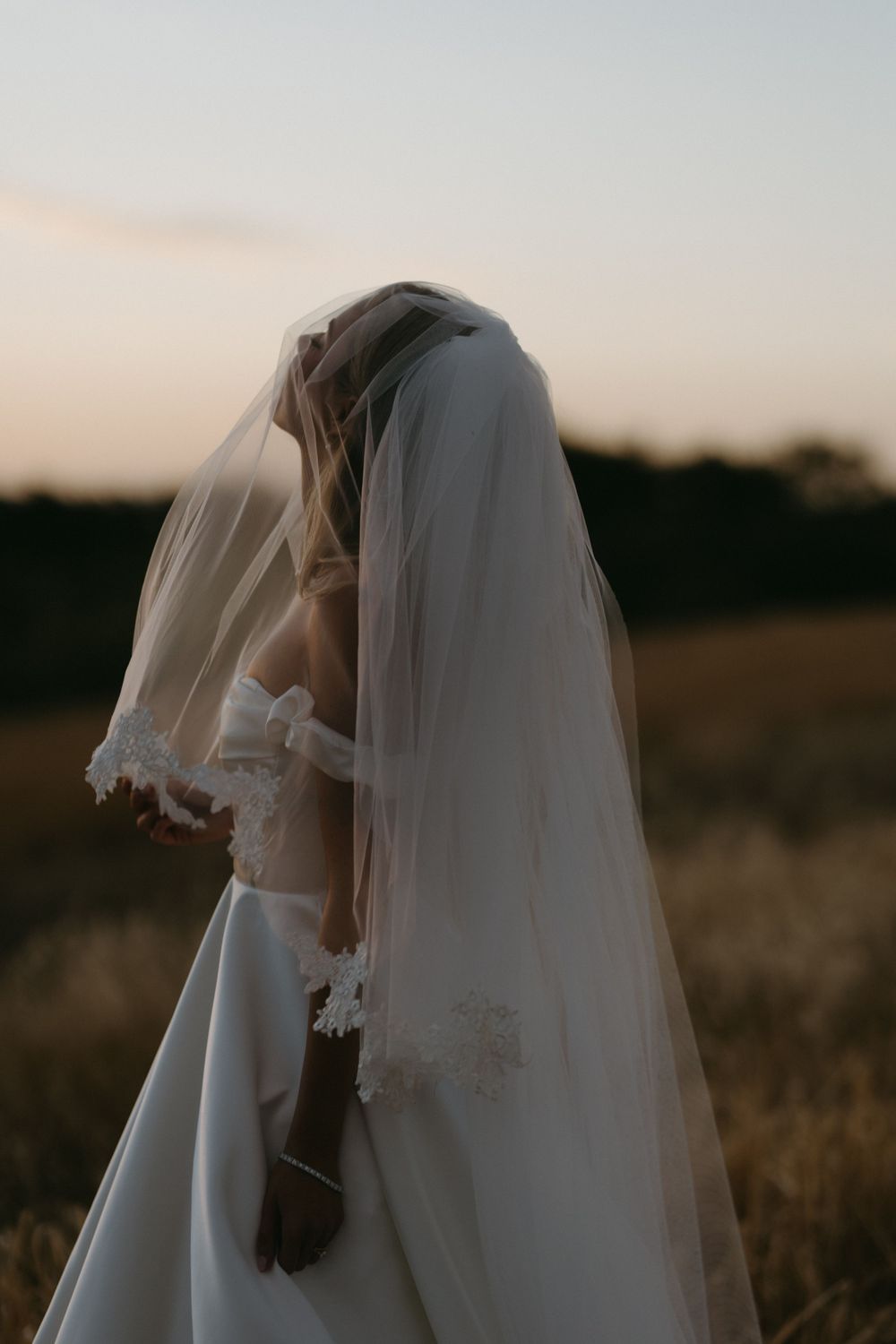 Ethereal side profile of a bride in a white dress with flowing veil against a dusky sky.