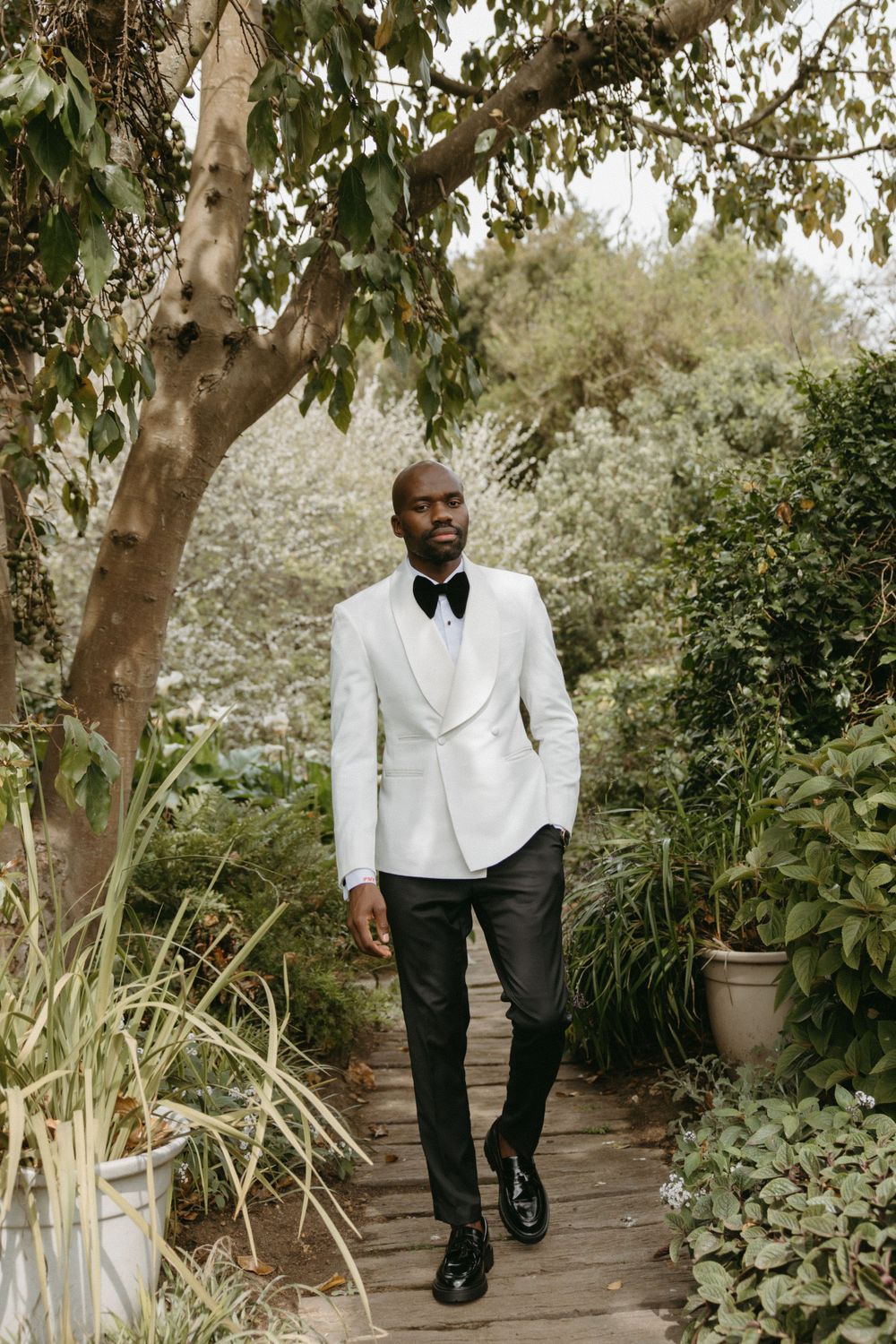 A groom in a white tuxedo walks along a garden pathway surrounded by lush greenery.