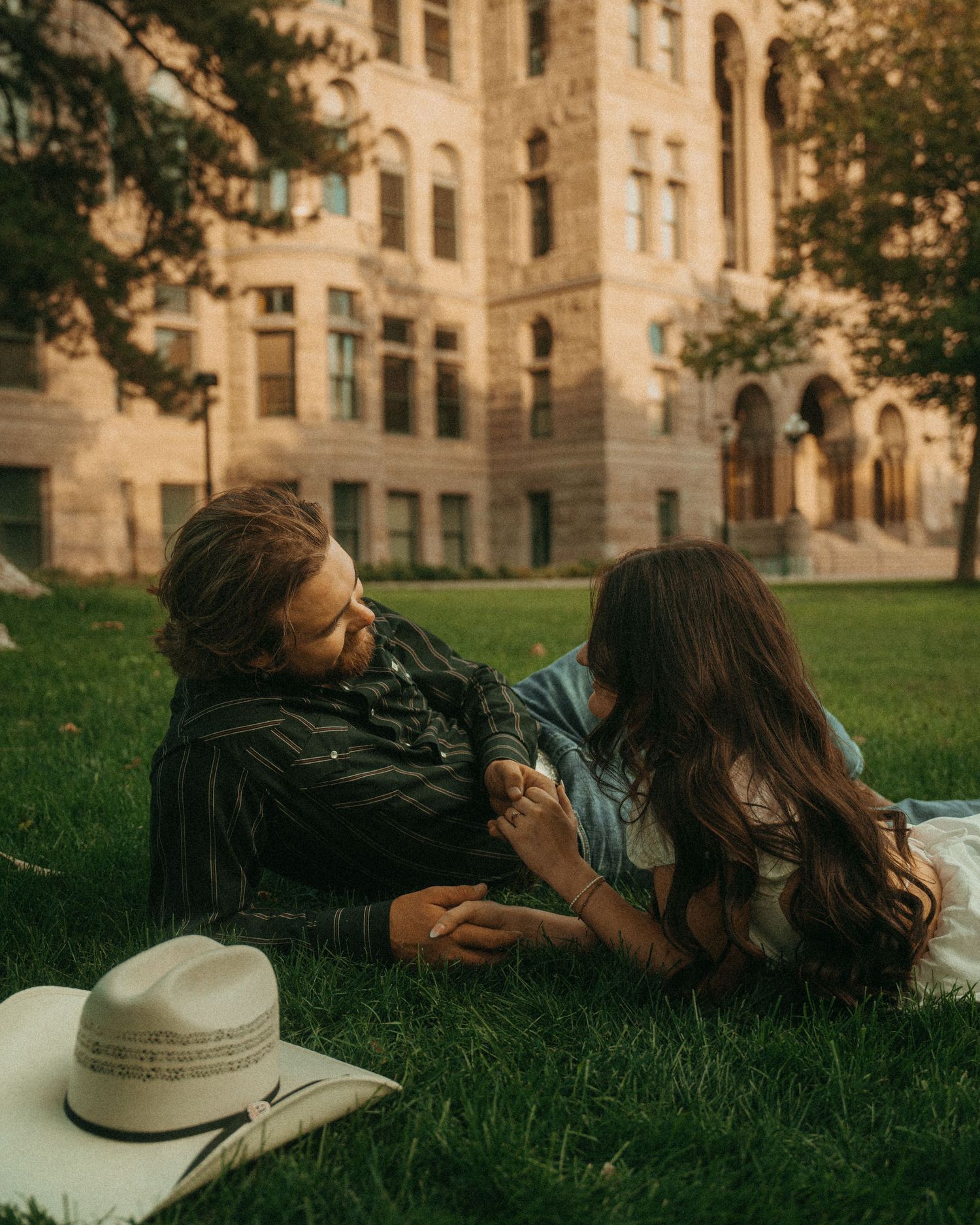 A couple lying on green grass near a historic stone building during golden hour with a hat resting nearby.