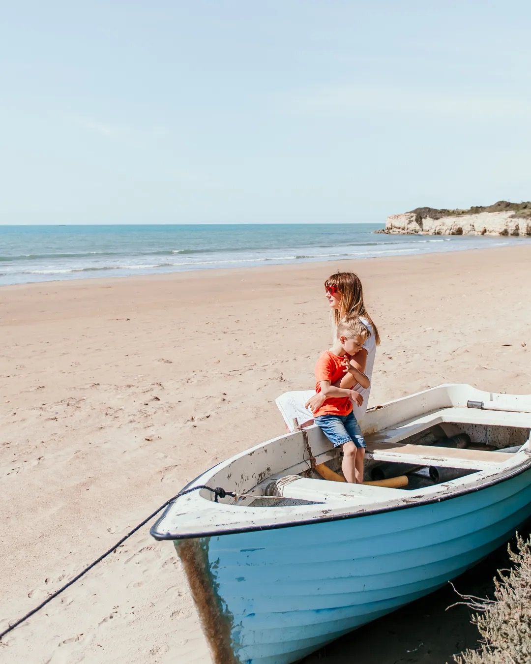 Young boy and mother sits in a small boat on a sandy beach, looking out toward the calm shoreline.