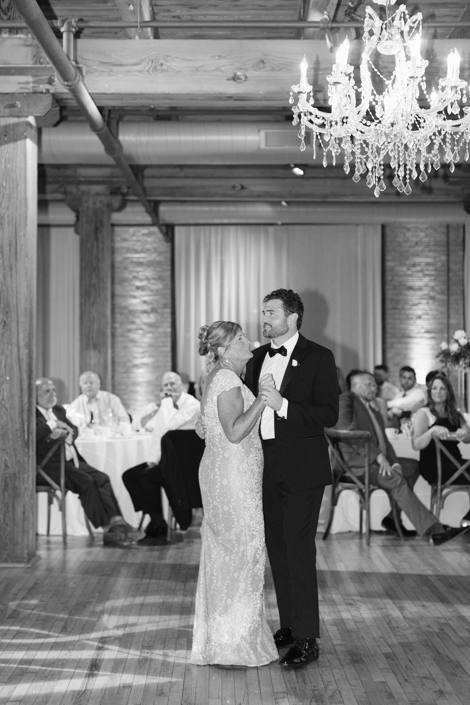 A couple shares their first dance in an elegant ballroom with wooden floors and crystal chandeliers overhead.