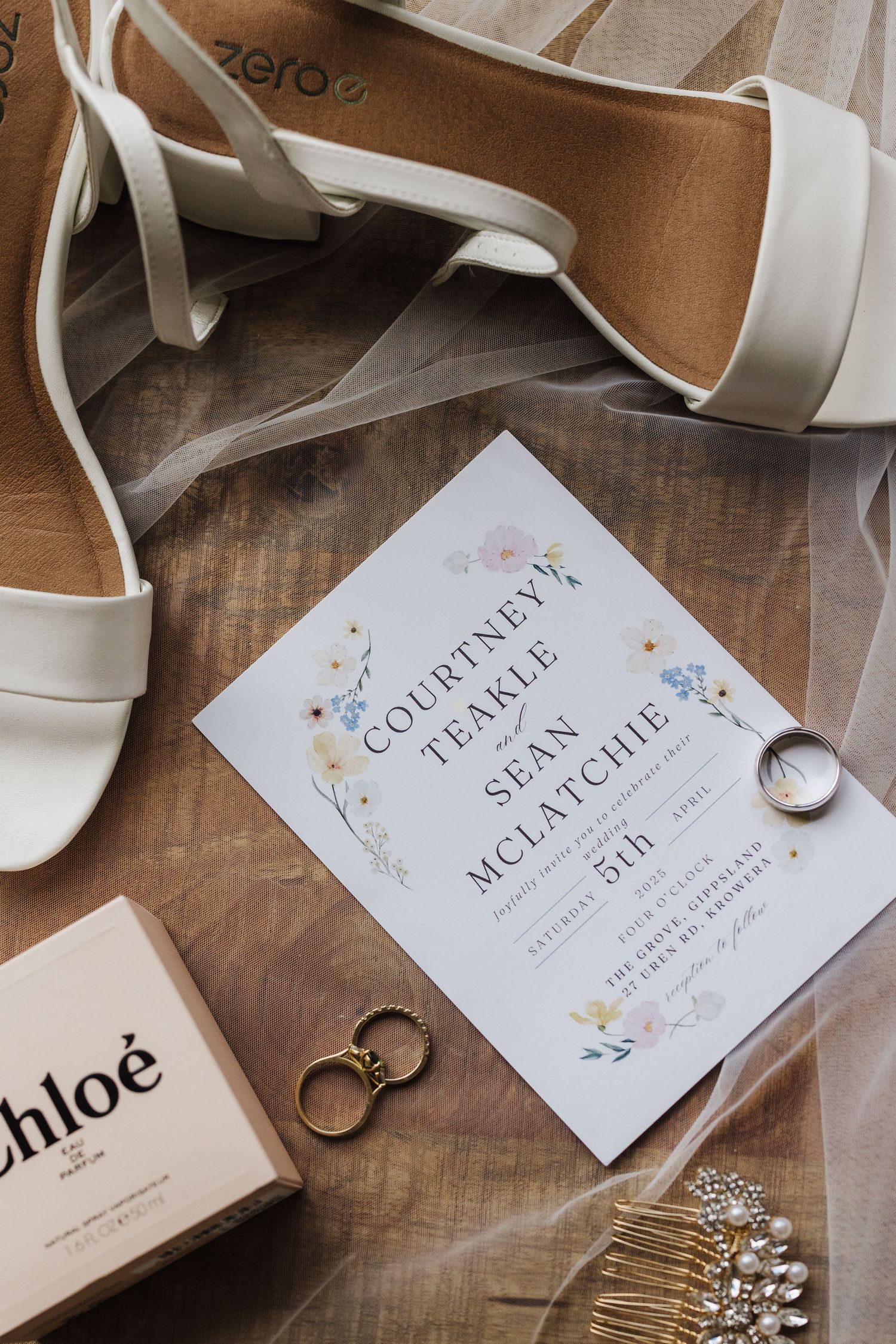 Wedding details flatlay with white heels, invitation card, gold rings, and hair accessories on wooden surface.
