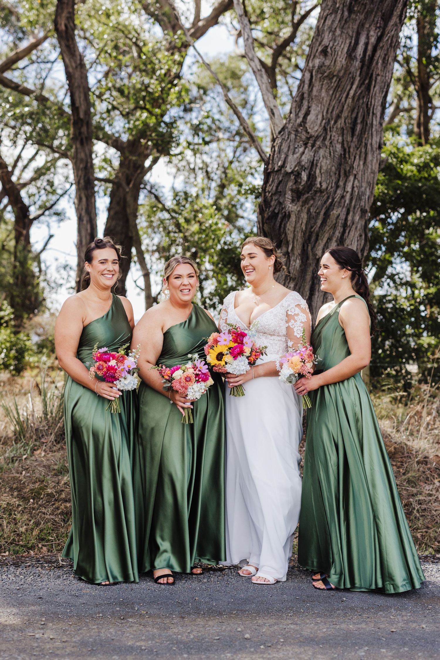 Wedding party in green satin bridesmaid dresses and white bridal gown holding colorful bouquets outdoors.