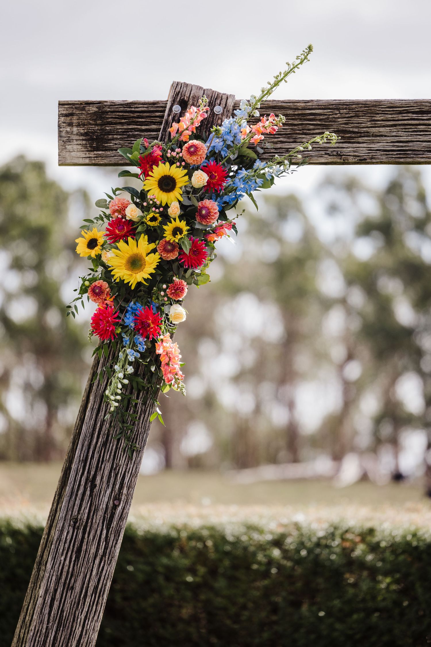 Rustic wooden fence post decorated with vibrant sunflowers, red roses, and blue wildflowers in a natural outdoor setting.
