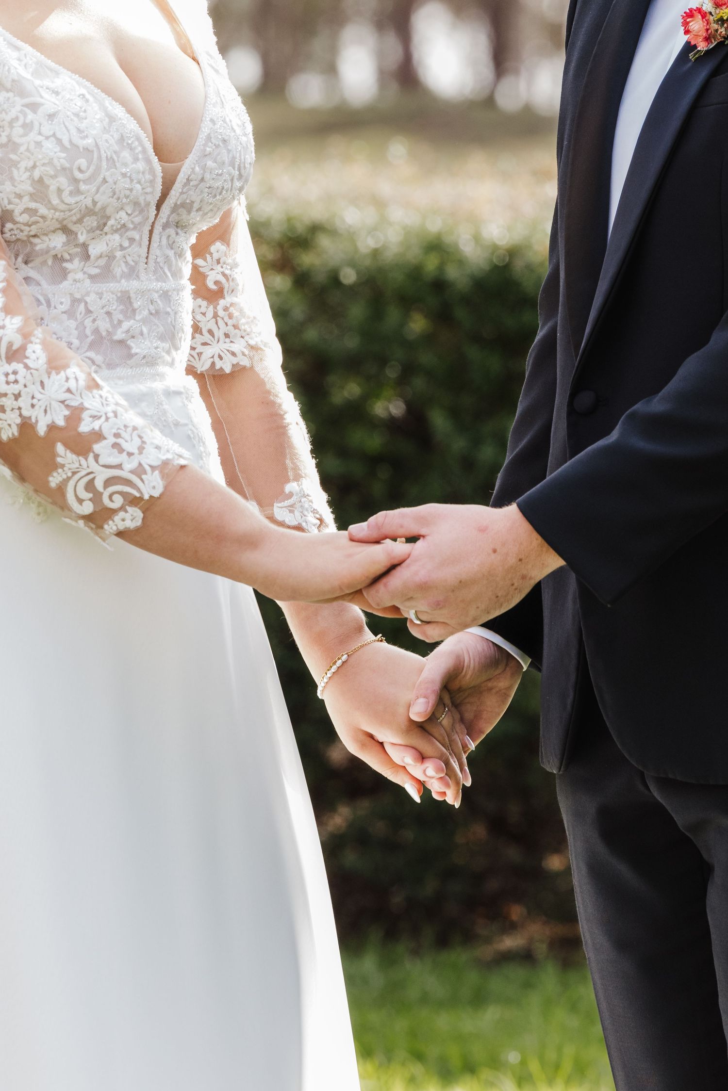 Close-up of couple holding hands at wedding ceremony, showing white lace dress sleeve and black suit jacket.