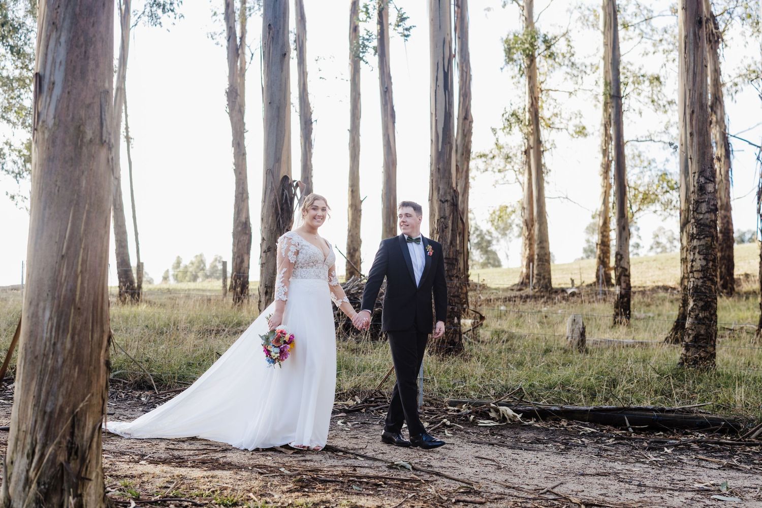 A couple in wedding attire stands among tall eucalyptus trees in a forest setting during a romantic photo session.