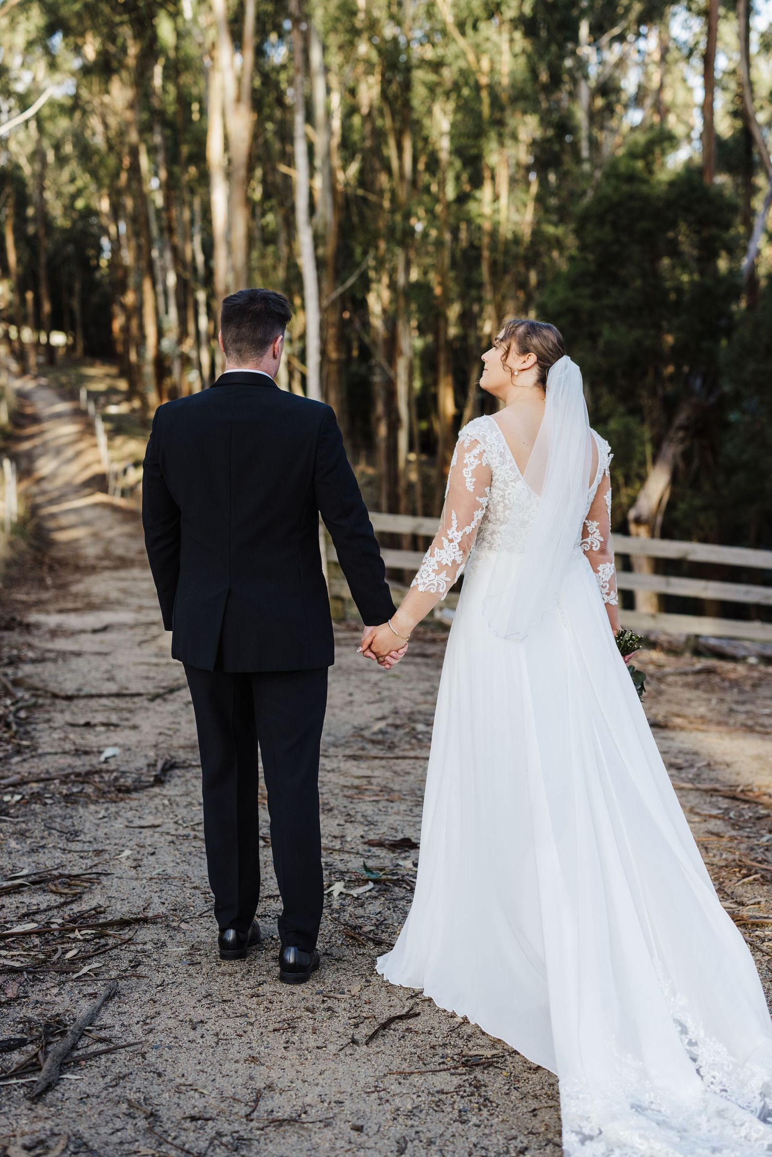A couple strolls hand in hand through a eucalyptus grove in wedding attire with a trailing lace veil.