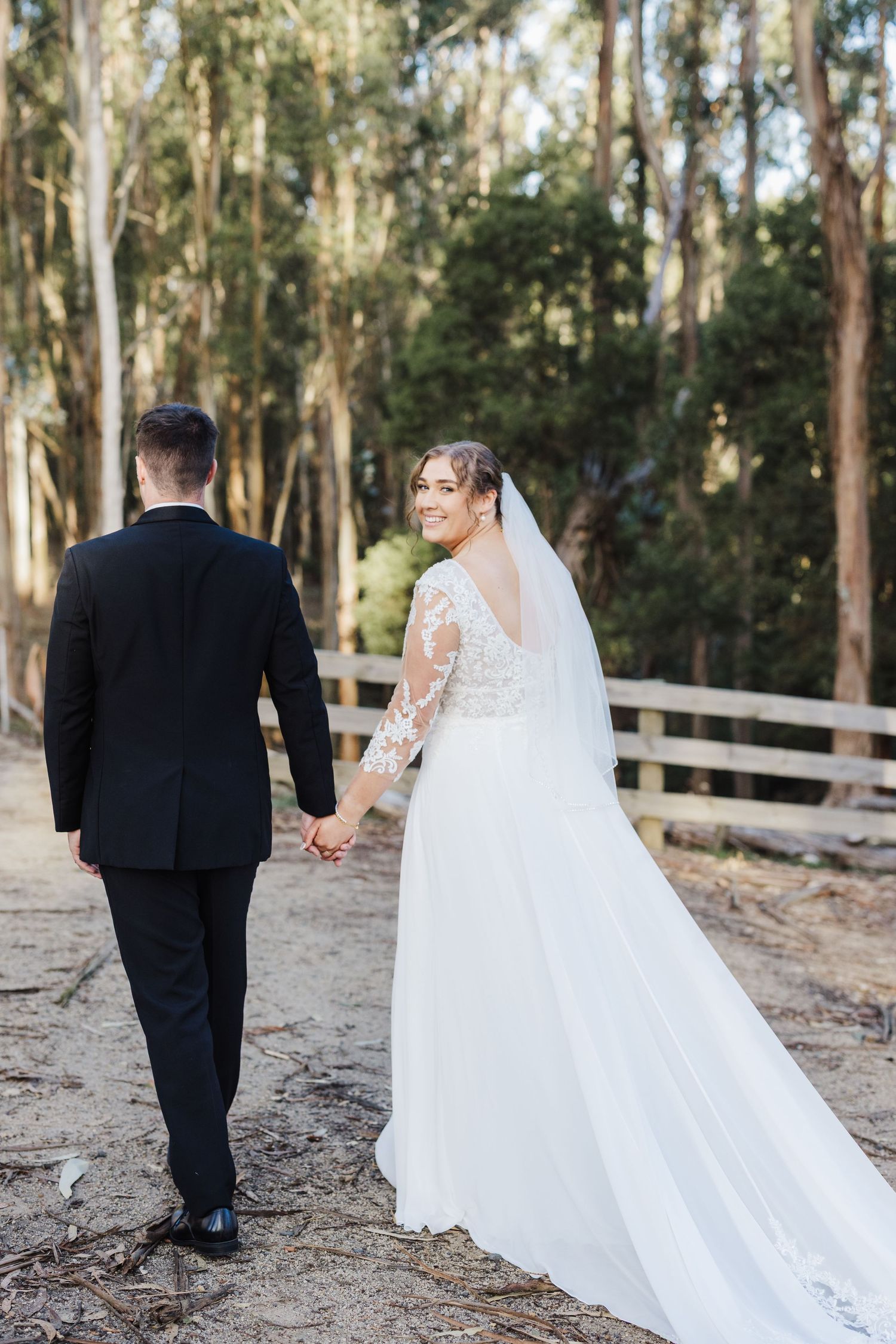 A couple walking along a wooded path in formal wedding attire with a long flowing white dress and black suit.