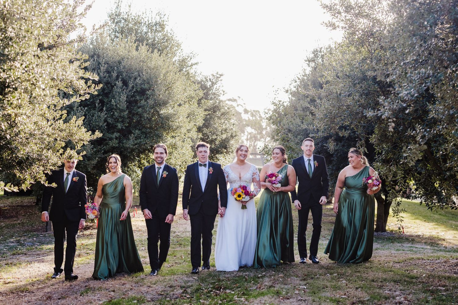 Wedding party in formal attire poses together on a tree-lined path with bridesmaids wearing emerald green dresses.