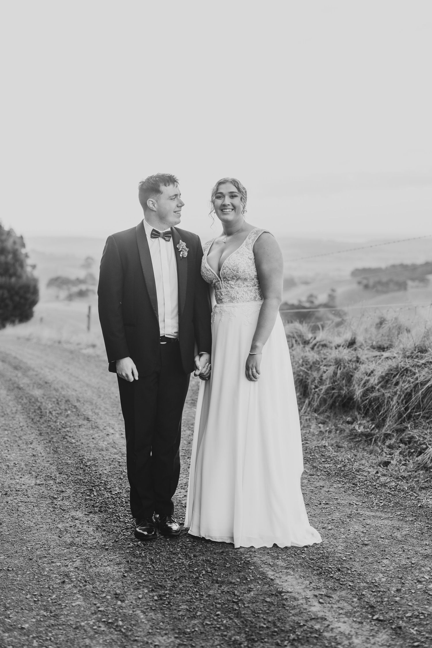 Black and white portrait of a couple in wedding attire standing on a rural dirt path.