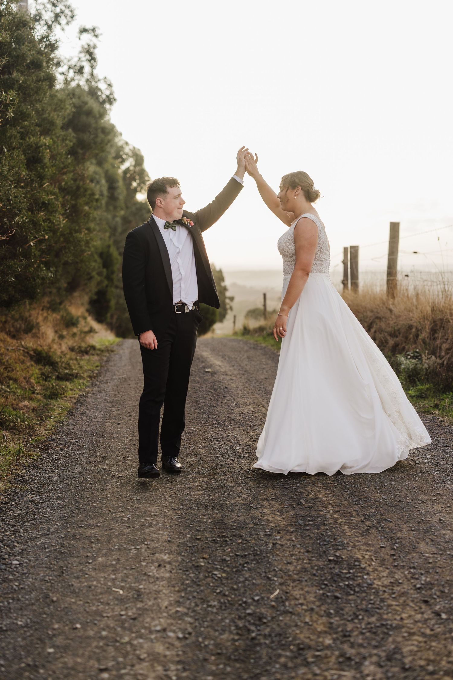 A couple in formal wedding attire share a playful moment on a gravel path at sunset.
