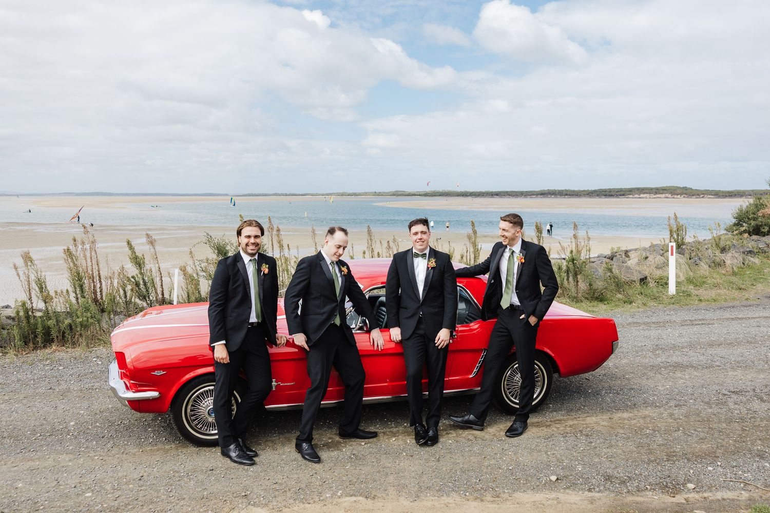 A group poses together by a vintage red convertible car on a scenic coastal road.