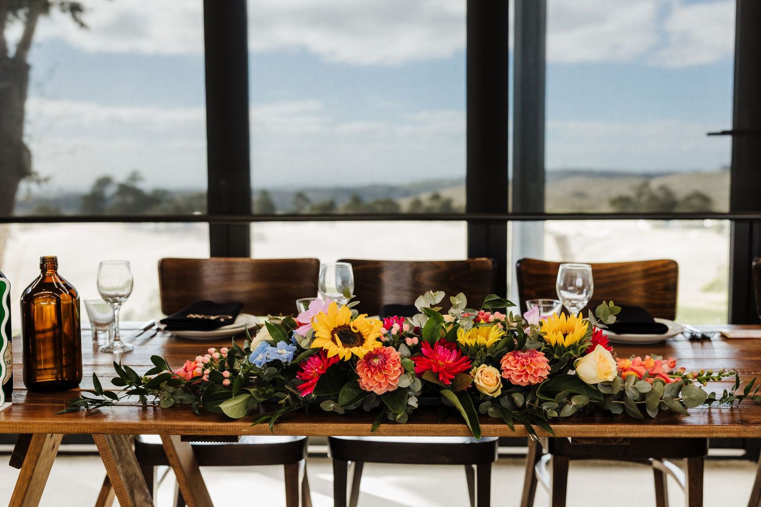 A long wooden dining table set with floral centerpieces and wine glasses against panoramic windows overlooking a landscape.