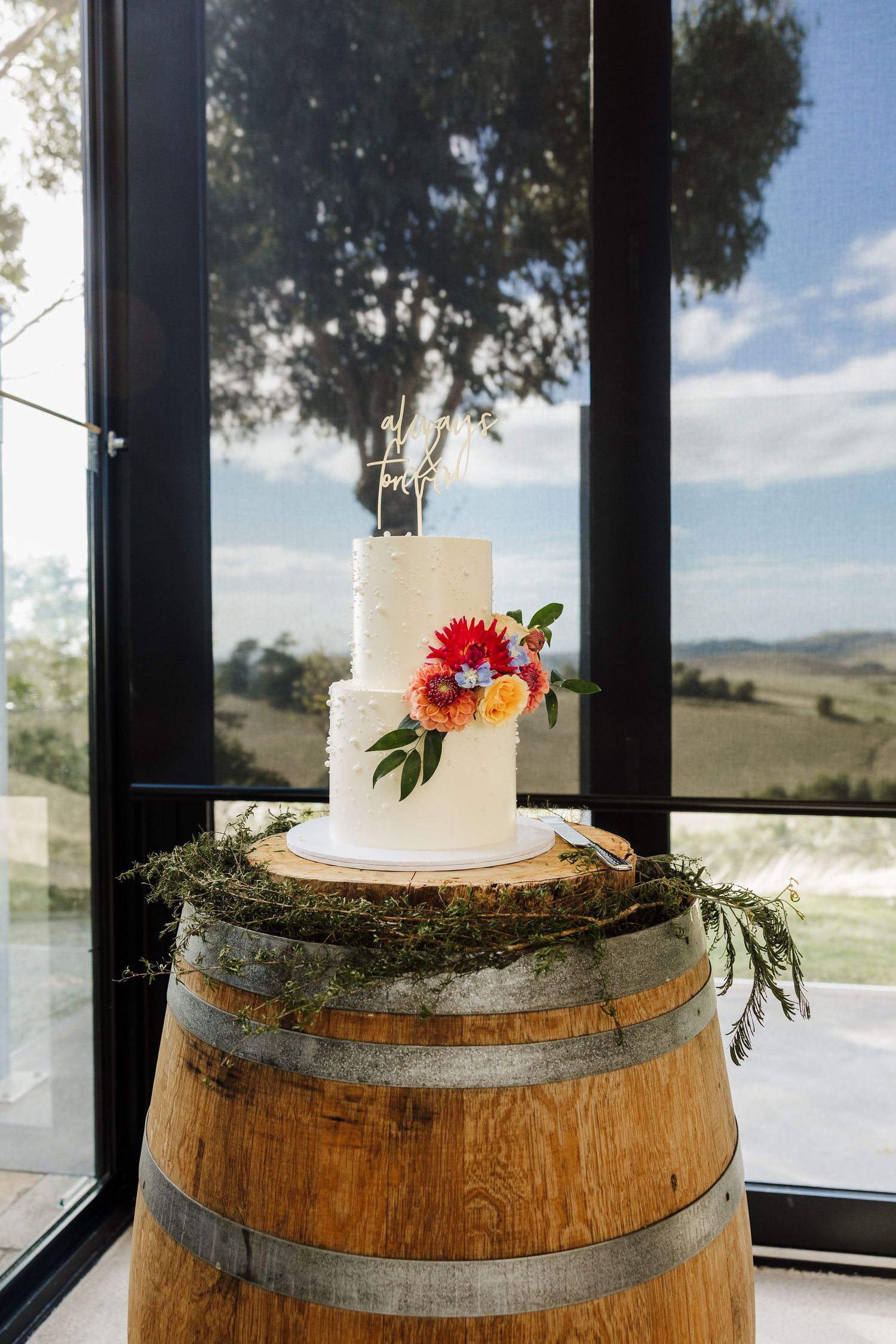 A white wedding cake adorned with colorful flowers sits atop a rustic wine barrel by a window overlooking rolling hills.