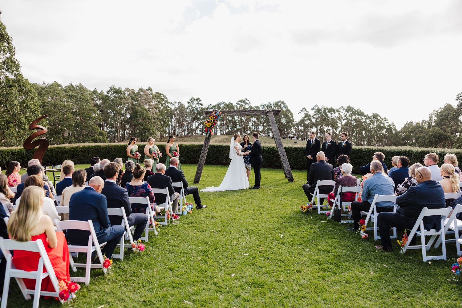 Outdoor wedding ceremony on a lush green lawn with white chairs and guests watching the couple exchange vows.