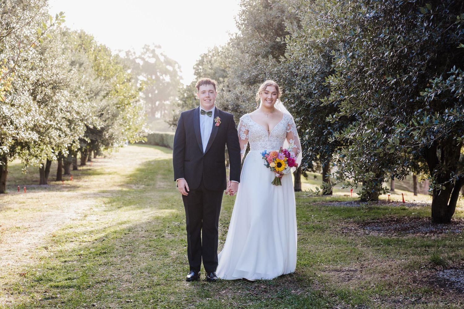 A couple stands together on a sunlit garden path surrounded by lush trees and greenery on their wedding day.