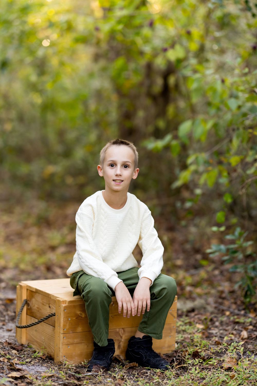 A child sits on a wooden crate in an autumn forest setting wearing a white sweater and olive pants.