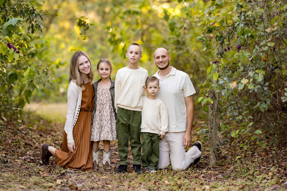 A family wearing coordinated white and brown outfits poses together in a natural outdoor setting with autumn foliage.