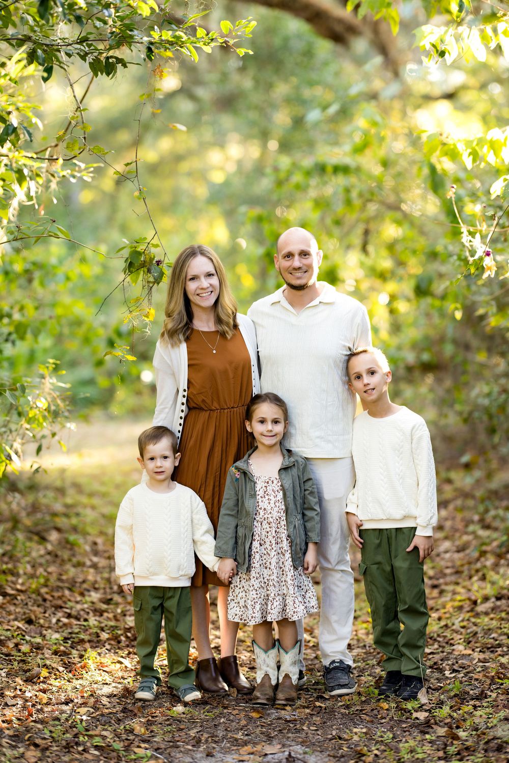 A family poses together outdoors in coordinated neutral outfits surrounded by sunlit autumn foliage.