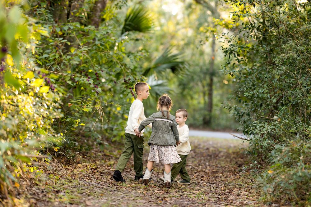 Two children walk hand in hand down a sunlit forest path surrounded by lush green foliage.