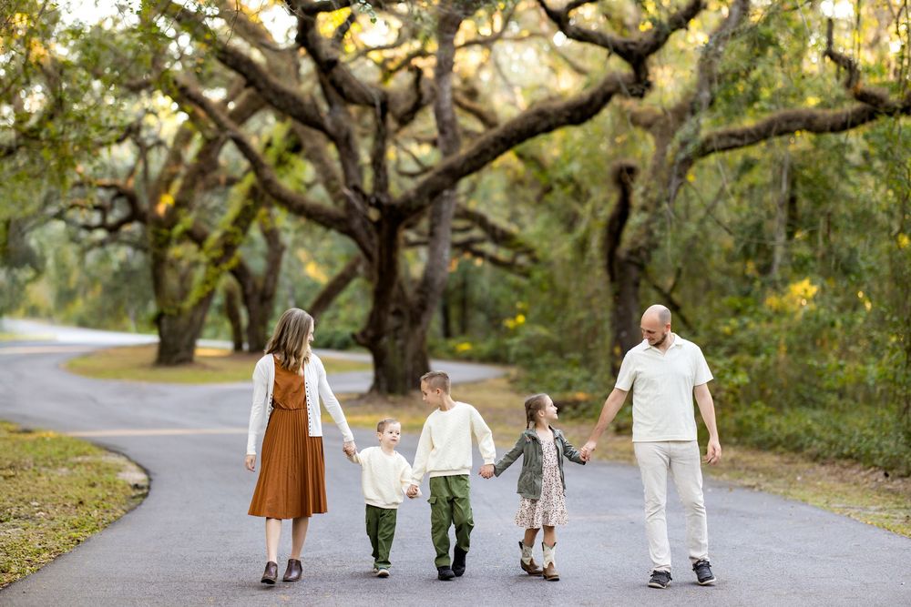 A family walks together holding hands along a winding path under majestic oak trees on an autumn day.