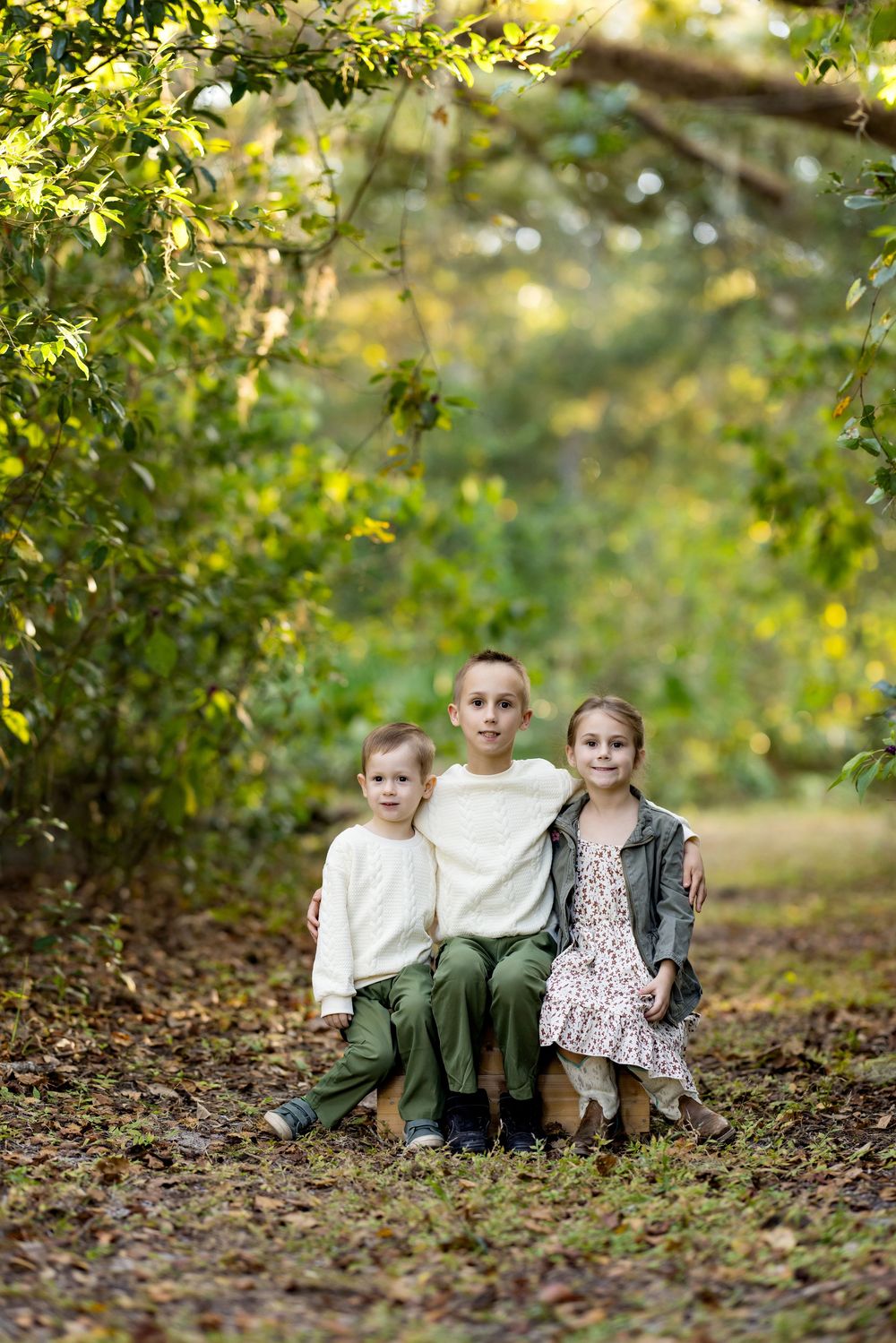 Two children sit together on fallen autumn leaves beneath green trees in a natural outdoor setting.
