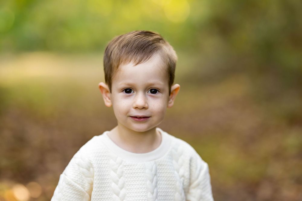 A toddler wearing a white knit sweater smiles while sitting outdoors in an autumn setting.