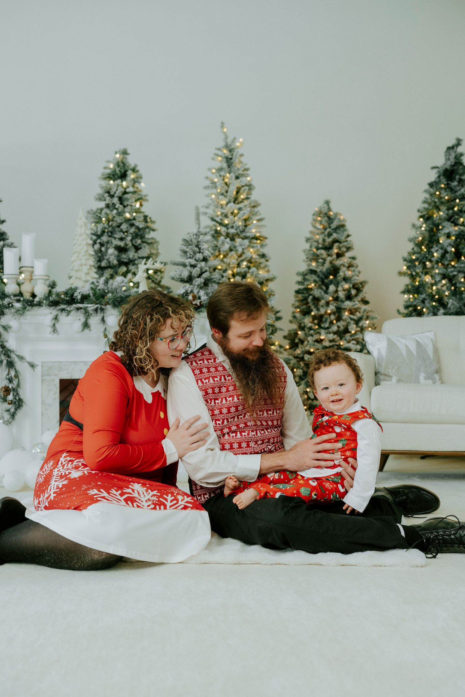 Family wearing matching red and white holiday outfits sits together on floor with decorated Christmas trees in background.