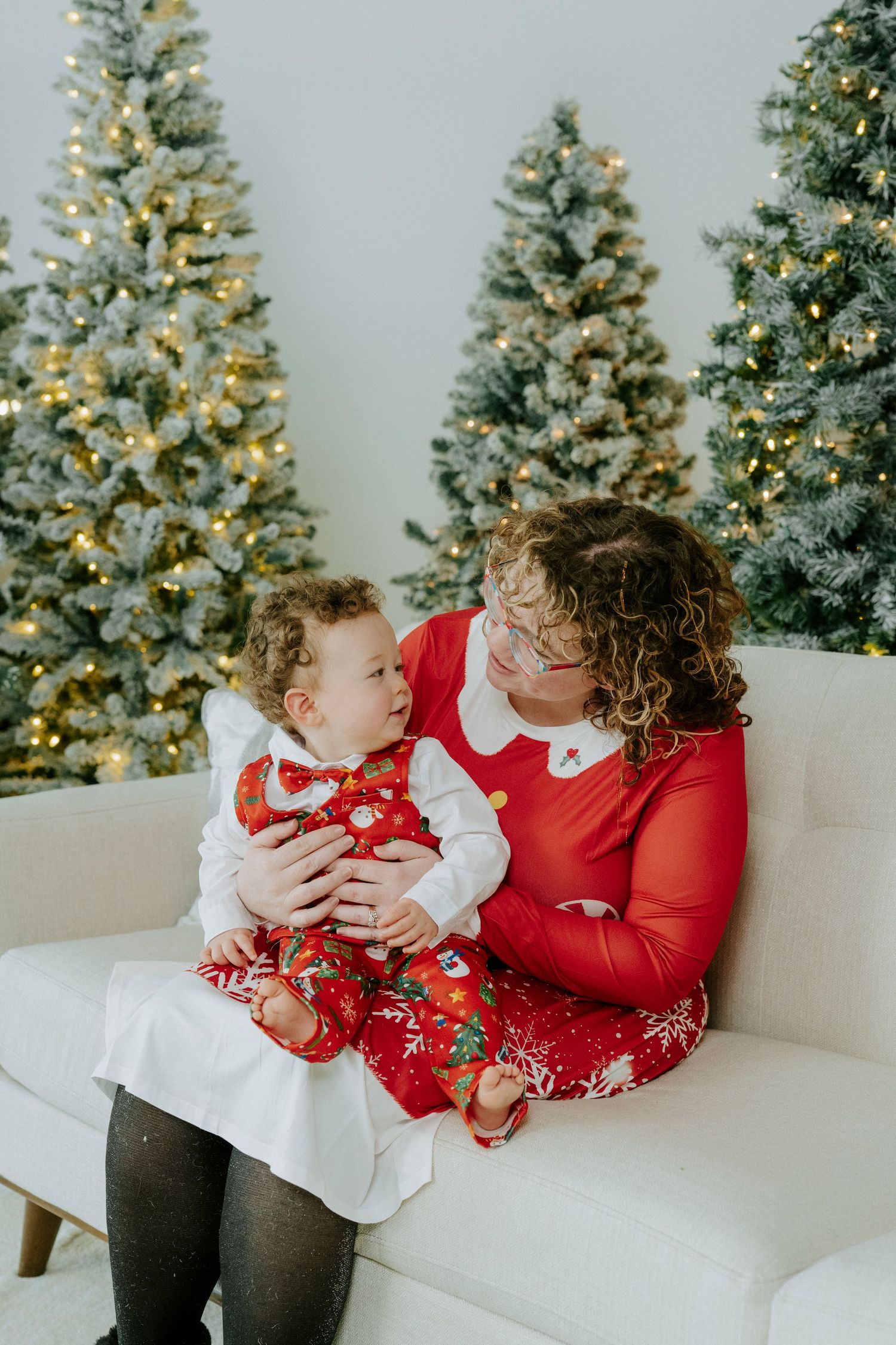 A heartwarming holiday scene with two people in red and white outfits sitting on a white couch near decorated Christmas trees.