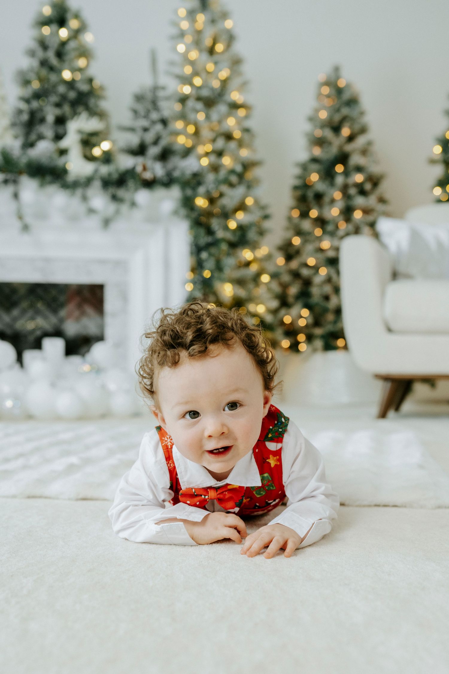 Baby in white and red holiday outfit lies on white carpet with lit Christmas trees in background.