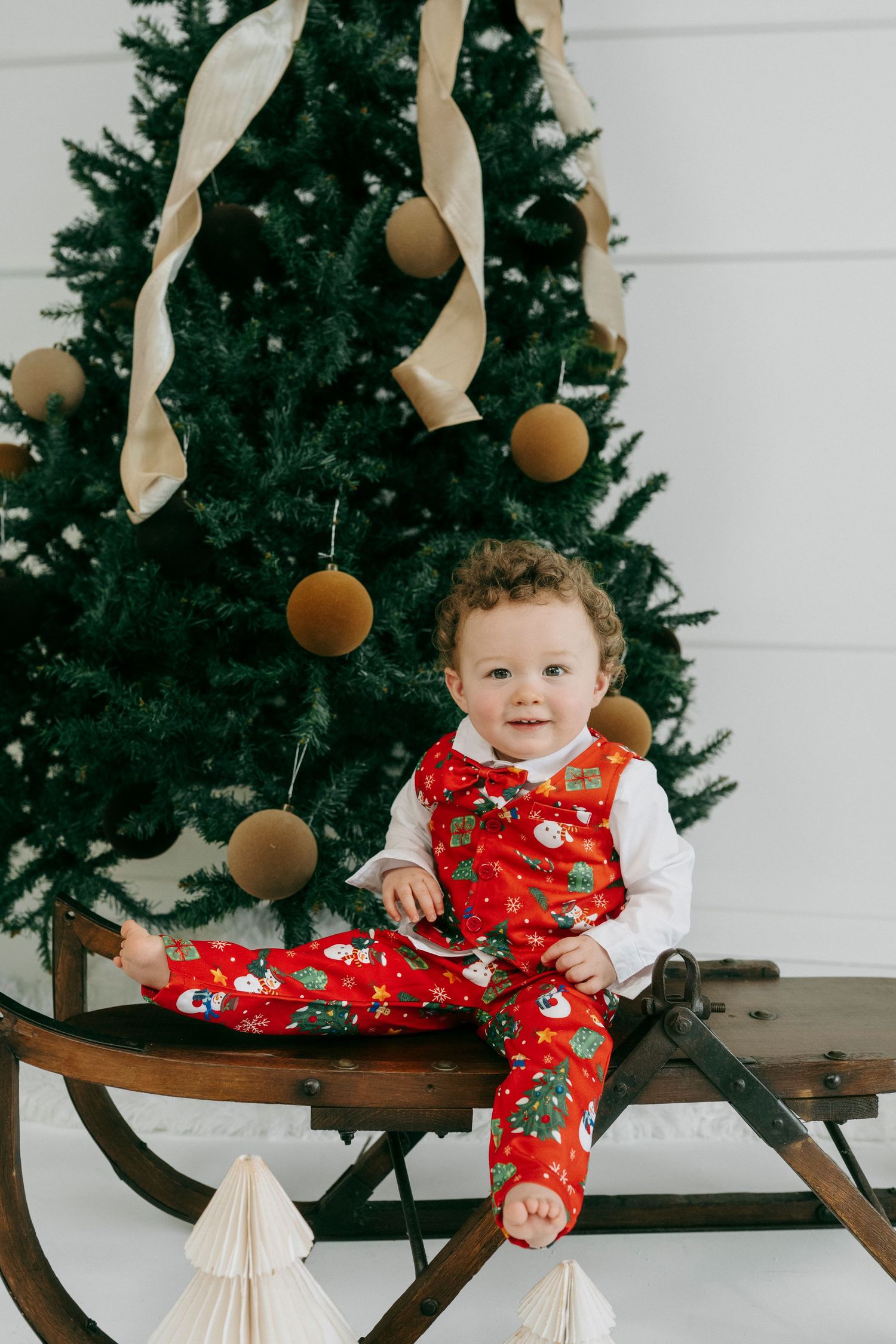 A decorated Christmas tree with gold ribbons and ornaments stands behind a vintage wooden sleigh with holiday decor.