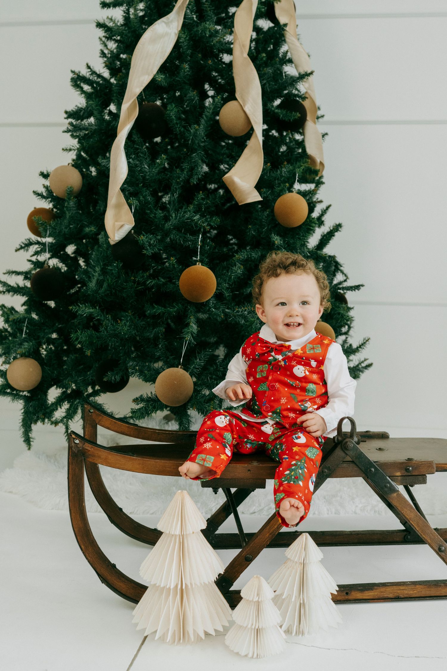 A baby in red holiday pajamas sits on a vintage wooden sleigh next to white paper Christmas trees.