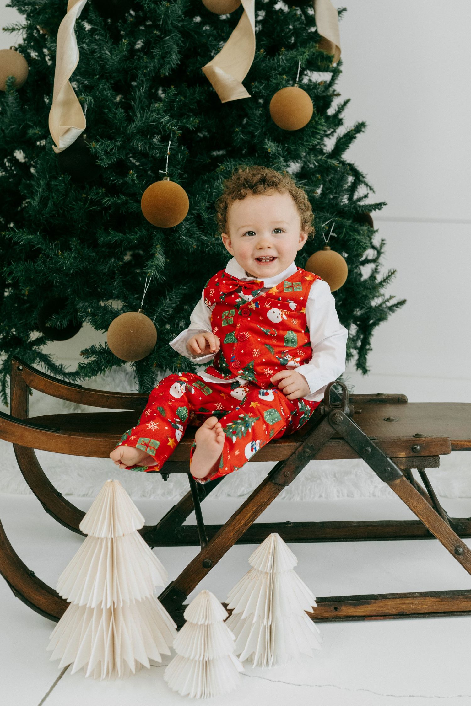 Child in red Christmas overalls sits on vintage wooden sleigh with paper tree decorations and ornaments on Christmas tree.