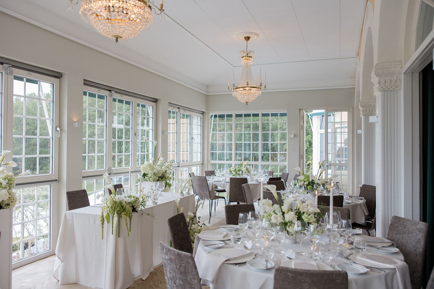 Elegant white wedding reception room with crystal chandeliers, round tables, and large windows overlooking garden views.