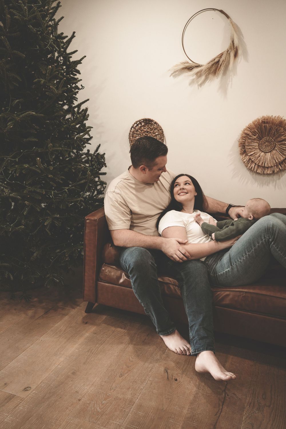 Couple lounges together on brown leather couch next to Christmas tree in cozy living room setting.