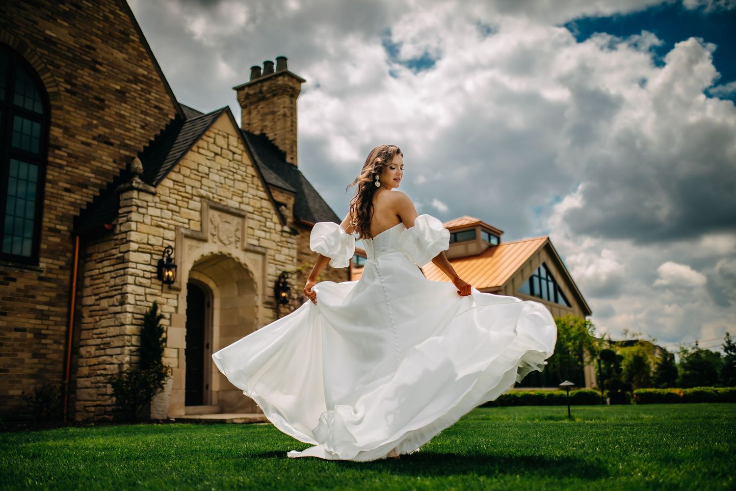 A bride in a flowing white dress twirls gracefully against a dramatic cloudy sky and stone church backdrop.